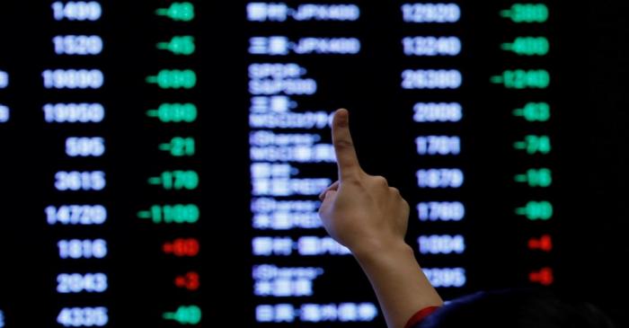 A woman points to an electronic board showing stock prices as she poses in front of the board