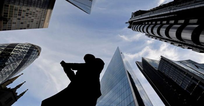 A worker looks at their phone as they walk past The Gherkin, Lloyds, and other office buildings