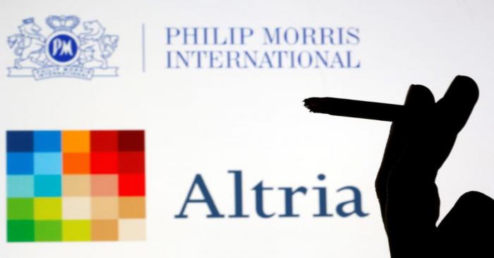 A hand with a cigarette is seen in front of displayed logos of Philip Morris and Altria in this