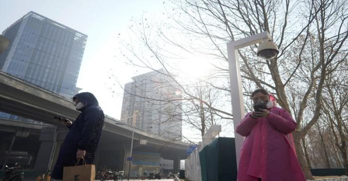 Pedestrians wearing masks stand under a surveillance camera as they wait to cross a road after