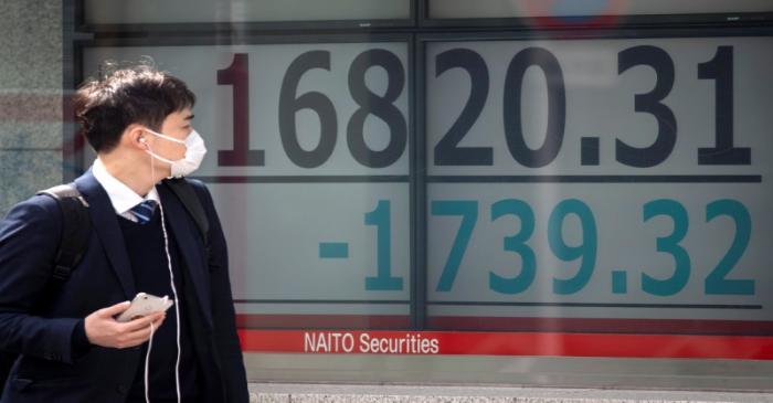 A man wearing a protective face mask walks past a screen showing Nikkei index in Tokyo