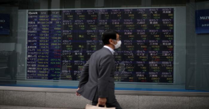 A man wearing protective face mask walks in front of a stock quotation board outside a