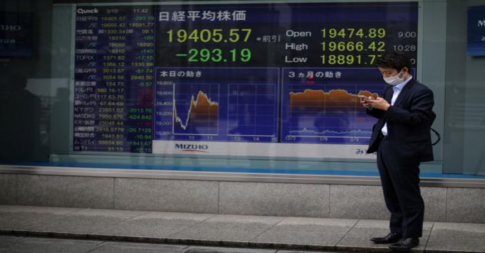 A man wearing protective face mask stands in front of a stock quotation board outside a