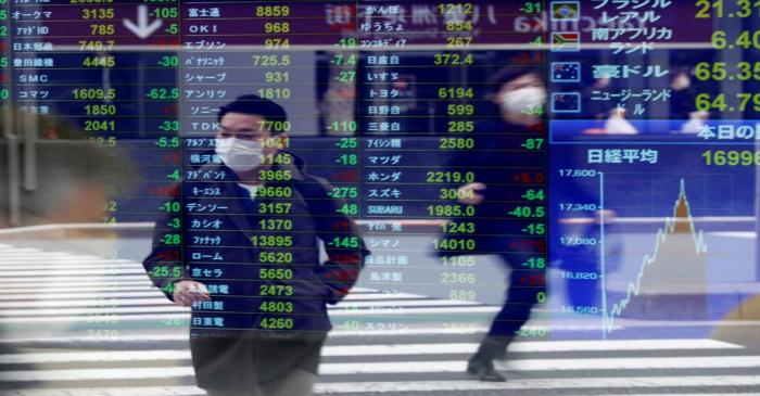 Passersby wearing protective face masks are reflected on a screen displaying stock prices