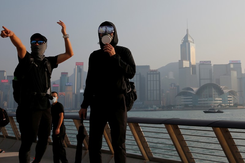 Masked anti-government protesters react in front of the skyline during a protest at Tsim Sha